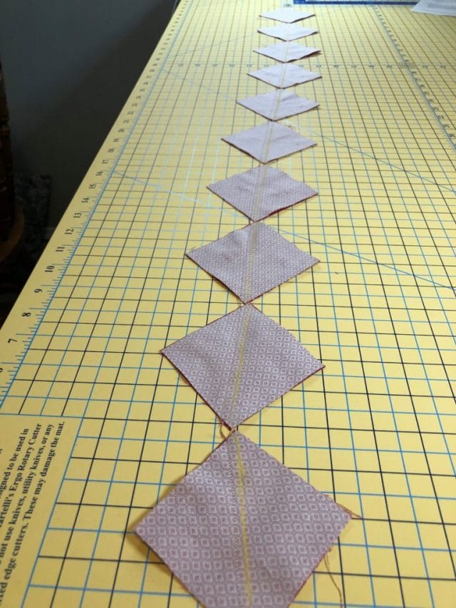 Squares of fabric with right sides facing have been sewn next to the marked lines into a strand and are resting on a cutting mat.