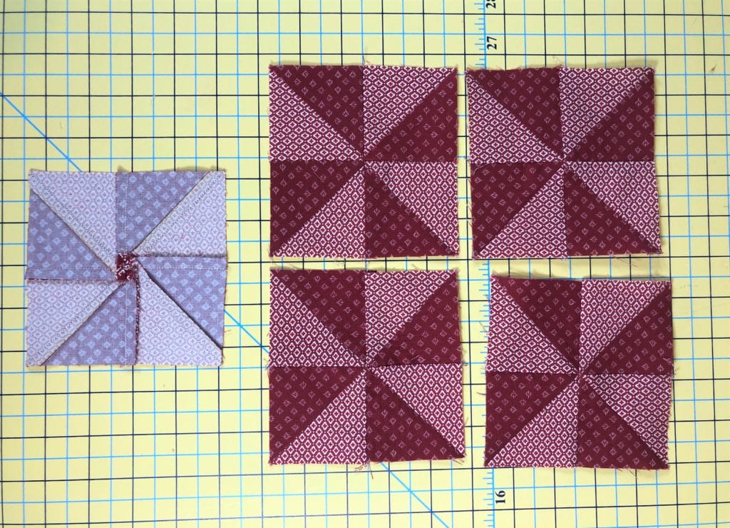 Red fabrics are sewn into 5 squares with a pinwheel design and are resting on a cutting mat.