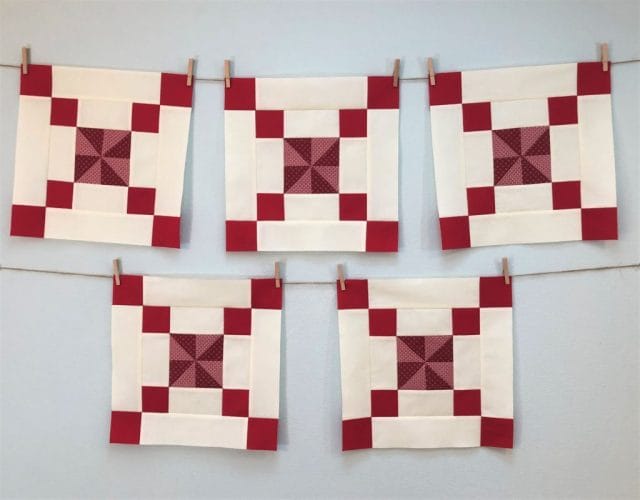 Five red and white quilt blocks hang from twine, they feature a red pinwheel design at center and small red squares at the corners.